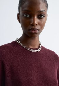 Burgunderfarbener Strickpullover mit rundem Ausschnitt, ausgestattet mit einer silbernen Halskette mit verlängerten Gliedern und strukturierten Akzenten.