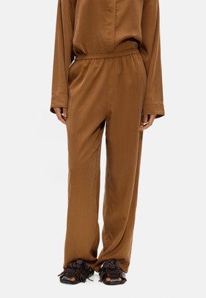 Osoba w luźnych brązowych spodniach i dopasowanym długim rękawem, stojąca, w ciemnobrązowych sandałach z frędzlami na białym tle.