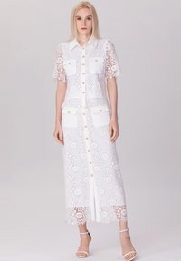 Robe en dentelle blanche à manches courtes, avec un devant à boutons, deux poches poitrine et un ourlet longueur sol. Présente une texture à motifs et un léger éclat.