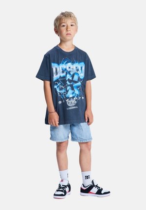 Dunkelblaue Graphic-T-Shirt mit einem Blitzdesign, kombiniert mit hellen Denim-Shorts. Schwarz-weiße Sneaker mit roten Akzenten runden das Outfit ab.