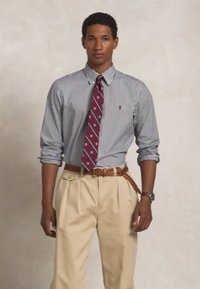 Ανδρικό πουκάμισο με κουμπιά σε μπλε και λευκές ρίγες, συνδυασμένο με μπεζ παντελόνια. Διαθέτει ένα μπορντό παπιγιόν με λευκές βούλες και μια πλεκτή ζώνη.