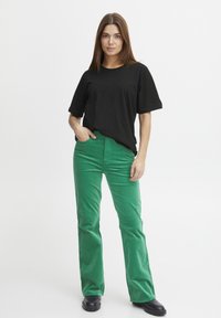 Sort, kortærmet t-shirt parret med grønne fløjl flared bukser og sorte støvler; model med hænderne på hoften, stående foran en hvid baggrund.