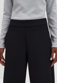 Zwarte wijde broek van soepel materiaal, met een platte tailleband en zijzakken, gecombineerd met een lichtgrijze lange mouwen top.