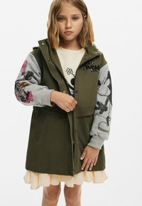 Olivgrön jacka med dragkedja, gråmönstrade ärmar och blommönster. Lager på en ljus, volangad krämfärgad klänning.