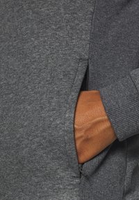 Grå sweatshirtdetalj som visar texturerad tyg, ribbade muddar och en hand som sträcker sig in i en dragkedjeficka.