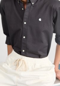 Camicia grigio scuro con bottoni e un piccolo logo bianco, abbinata a pantaloni color crema con coulisse, realizzati in tessuto strutturato e dalla vestibilità comoda.