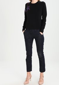 Schwarzer Strickpullover mit rotem und blauen Sternestick auf der Schulter, kombiniert mit dunkelblauen Jeans und nude-farbenen, geschnallten High Heels.