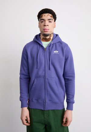 Felpa zip-up viola in un materiale morbido con logo frontale, cappuccio con cordino e tasche a marsupio; indossata con pantaloni verdi.