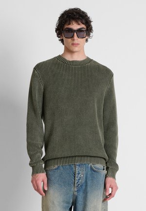 Giovane uomo con maglione a coste verde scuro, jeans blu scoloriti e grandi occhiali da sole neri, in piedi davanti a uno sfondo semplice.