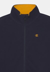 Tmavě modrá fleecová bunda s vysokým límcem, se zářivě žlutým vnitřkem a malým žlutým logem na hrudi. Přední zapínání na zip.