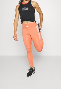 Zwarte mouwloze top met "Nike" tekst, oranje hooggetailleerde leggings met een zijpatroon en zwarte sneakers, in een studio-omgeving.