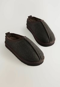Sötétbarna, slip-on papucs pár herringbone mintázatú anyaggal, puha belső béléssel és vastag gumitalppal egy fehér felületen.
