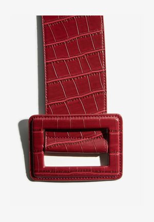 Široký červený opasek s krokodýlí texturou a velkou obdélníkovou sponou, vyfotografovaný na bílém pozadí se stínem.