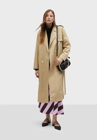 Beige double-breasted trench coat with gold buttons, worn over a black turtleneck and a pink striped skirt. Includes a black handbag and flat shoes.