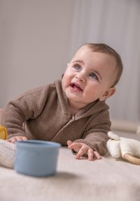 Brauner Strickhoodie, blauer Plastikbecher und gestricktes Spielzeug auf neutralem Untergrund. Das Baby hat blaue Augen und kurze braune Haare und greift nach vorne.