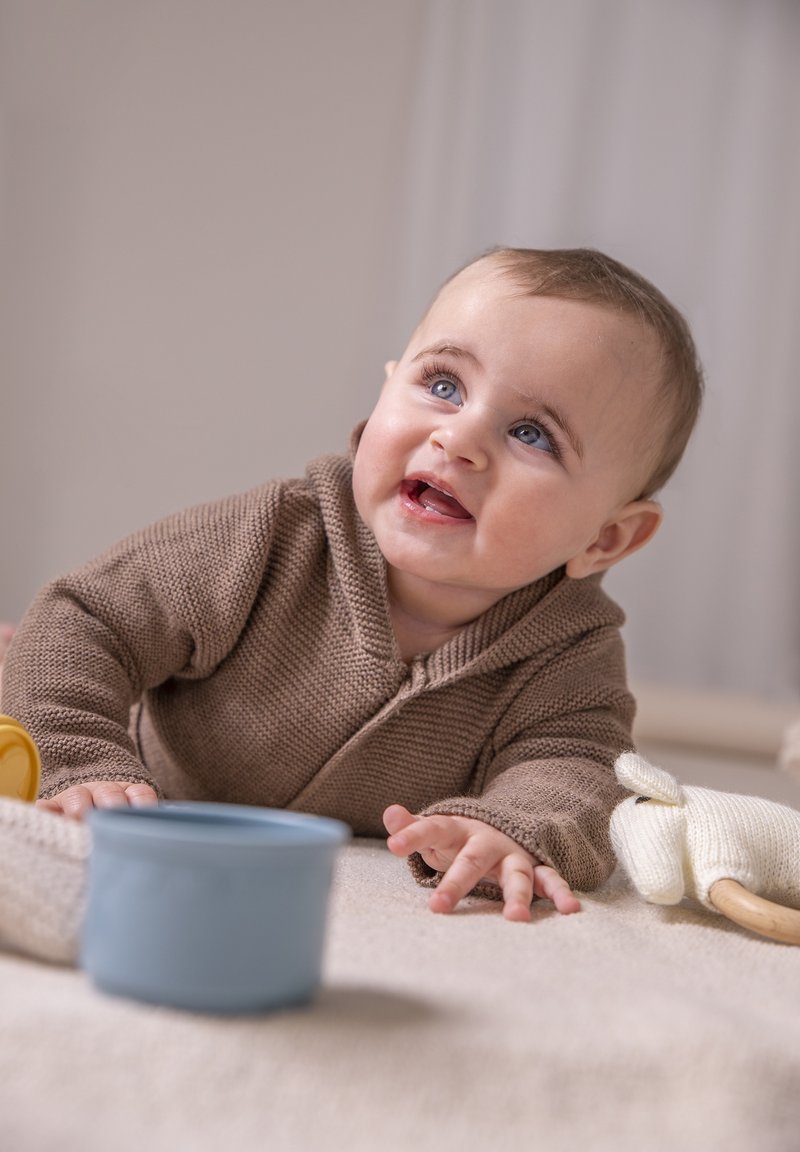 Brauner Strickhoodie, blauer Plastikbecher und gestricktes Spielzeug auf neutralem Untergrund. Das Baby hat blaue Augen und kurze braune Haare und greift nach vorne.