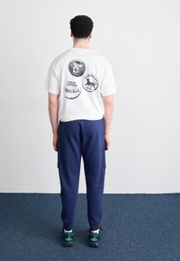 Man som står med ryggen mot kameran, iförd en vit T-shirt med tre svarta cirkulära logotyper och marinblå byxor, ståendes på en blå mattgolv.