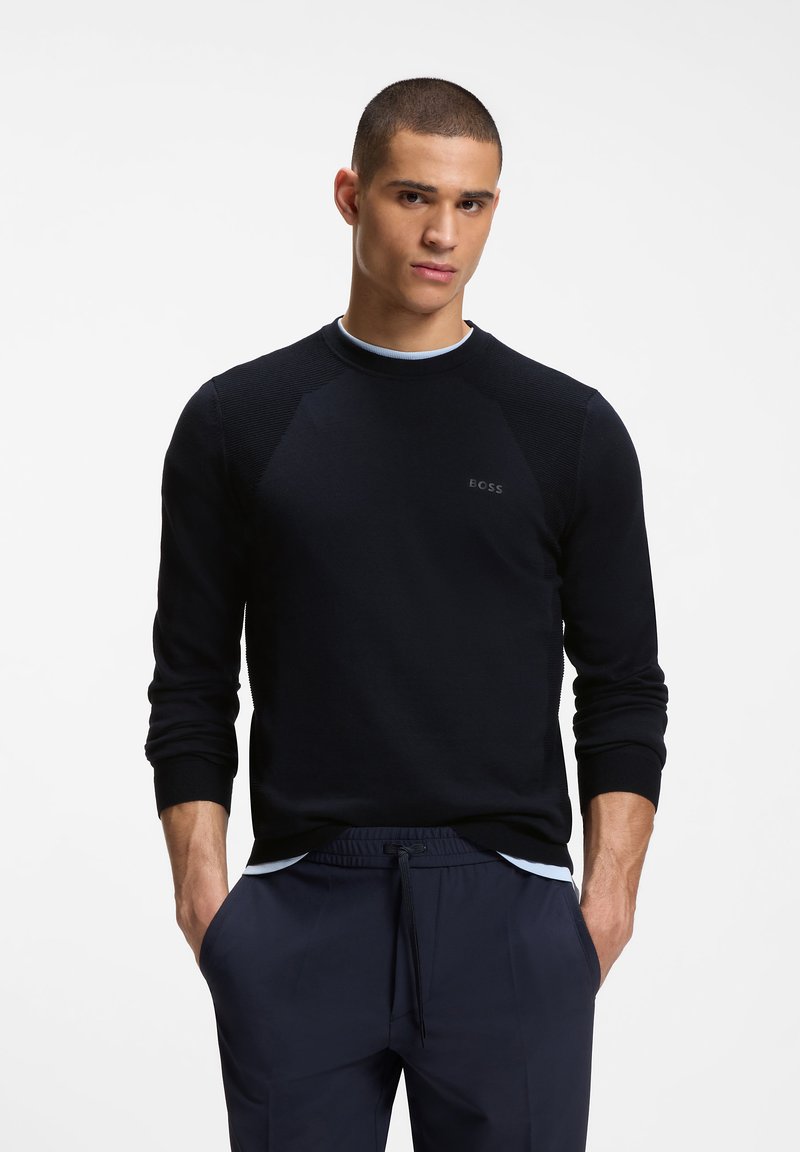 Νέος άνδρας με κοντά μαλλιά που φοράει μαύρο μακρυμάνικο πουκάμισο και ναυτικό μπλε παντελόνι με κορδόνι, στέκεται με τα χέρια στις τσέπες.