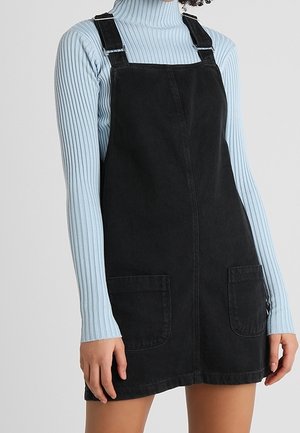 Frau trägt ein schwarzes Denim-Latzkleid mit vorderen Taschen über einem hellblauen gerippten Rollkragenpullover.