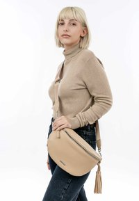 Beige leren schoudertas met een gestructureerde afwerking, ritssluiting en een kwastaccent. Draagt aan de linkerkant over een tan trui en jeans.