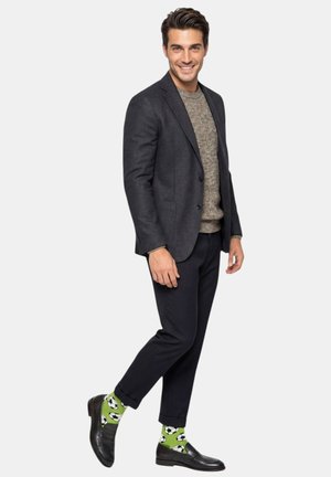 Lächelnder Mann in schwarzem Blazer, grauem Pullover, schwarzer Hose, schwarzen Schuhen und grünen Socken mit schwarz-weißem Fußballmuster, der auf einem Bein steht.