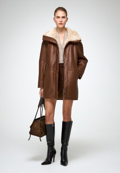 Bruine leren jas met een bontkraag, knielange ontwerp, gecombineerd met een beige top, een bruine rok en zwarte kniehoog stiletto laarzen.