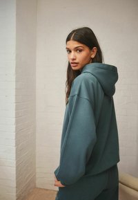 Teal übergroßer Hoodie mit einer Fronttasche und Kapuze mit Kordelzug, aus weichem Material gefertigt, mit entspannten Ärmeln und strukturierter Oberfläche.
