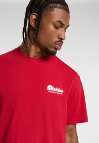 Camiseta roja de manga corta hecha de algodón. Presenta un logo blanco que dice "Dickies Workwear Company" en el área del pecho izquierdo. Diseño sencillo.