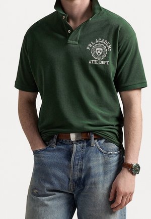 Mand iført mørkegrøn polo med logoet "PRL Academy Athl Dept.", lyseblå jeans, brunt bælte og brun læderur.