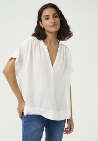 White short-sleeve blouse with a V-neck and gathered shoulders, made of lightweight fabric. Worn over blue denim jeans.