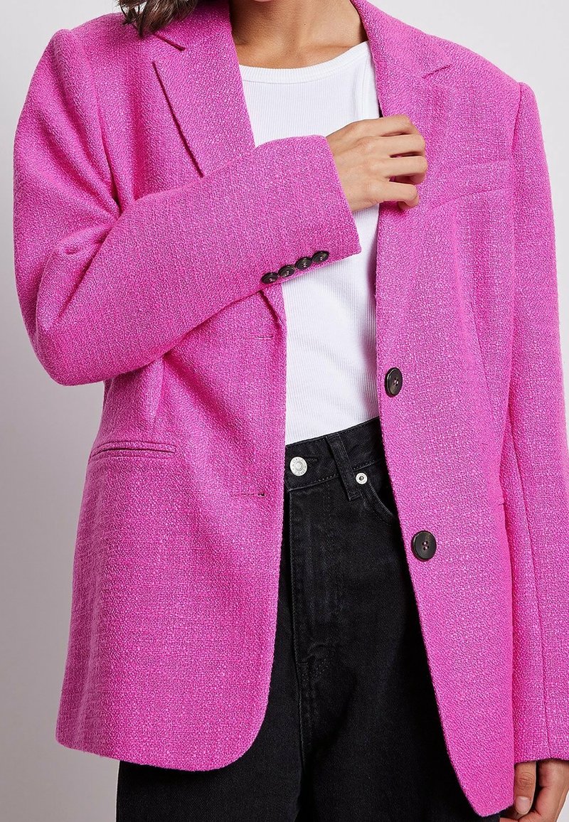 Femme ajustant un blazer rose vif texturé porté sur une chemise blanche et un pantalon noir, vue rapprochée des épaules aux hanches.