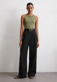 Top sans manches vert olive, pantalon noir taille haute à jambes larges, ceinture noire à boucle, sandales plates noires, sur un fond neutre.
