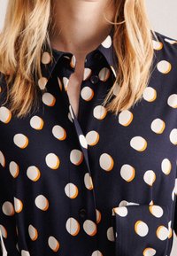 Blusa de color marino con un patrón de lunares en blanco y naranja, que presenta un cierre de botones en la parte delantera, cuello y bolsillo en el pecho. Textura de tela suave.