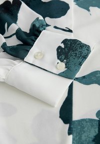 Witte blouse stof met een groenblauw abstract bloemenpatroon, met een geplooide manchette voorzien van twee crèmekleurige knopen bij de pols.
