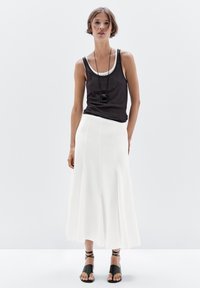 Femme portant un haut sans manches noir, une longue jupe blanche, des sandales noires à lacer et un collier pendentif noir, se tenant contre un fond blanc.