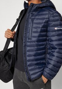 Person wearing a navy blue puffer jacket with Champion logo, black shirt, gray pants, holding black gym bag strap over shoulder.