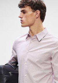 Lichtroze gestreepte shirt met een button-down kraag, met verticale strepen en subtiele textuur. Model houdt een zwarte tas vast.
