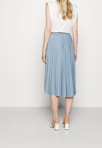 Hellblauer Faltenrock mit schimmerndem Bund, aus einem fließenden Material. Kombiniert mit weißen Sandalen mit klobigem Absatz.