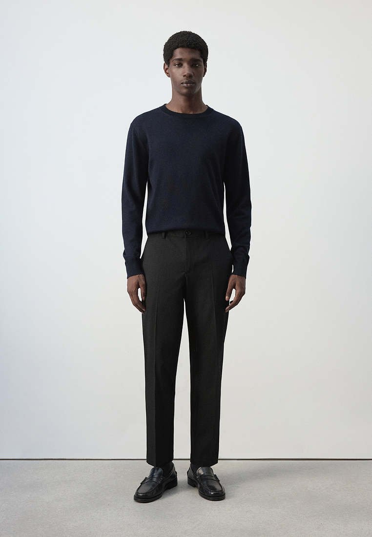 Homme debout portant un pull bleu marine foncé à manches longues, un pantalon noir sur mesure et des mocassins en cuir noirs sur un fond clair uni.