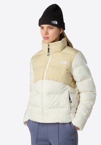 Ung kvinna som bär en beige och vit North Face pufferjacka, svart mössa och blå byxor med händerna i fickorna, som vänder sig något åt höger.