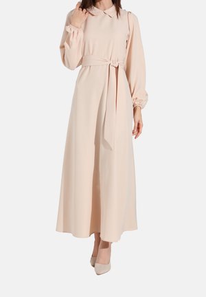 Frau trägt ein langes, blassrosa Kleid mit gebundenem Gürtel, langen Ärmeln und Kragen, kombiniert mit passenden blassrosa Pumps.
