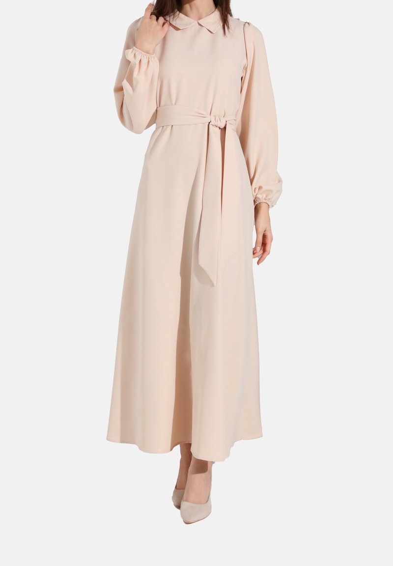 Femme portant une longue robe rose pâle avec une ceinture nouée, des manches longues et un col, associée à des talons rose pâle assortis.