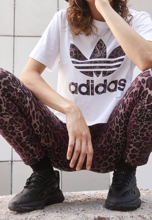 Personen sidder i squat iført en hvid Adidas t-shirt med leopardprint-logo, matchende leopardprintbukser, sorte sneakers og sølvhalskæder.
