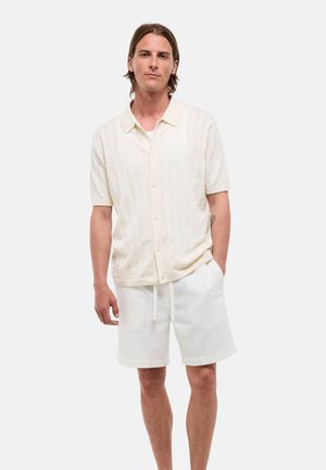 Homme aux cheveux mi-longs portant une chemise crème à manches courtes boutonnée et un short assorti, debout avec une main dans la poche.