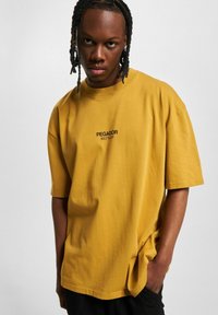 Gul bomulls oversized T-skjorte med rund hals og kort erm. Har svart trykk av "PEGADOR BUILT TO LAST" på brystet.