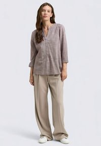 Világoslila, texturált tunika V-nyakkal és háromnegyedes ujjakkal, laza bézs nadrággal és fehér sportcipővel párosítva.