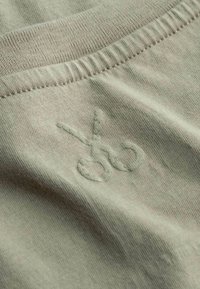 Tessuto di cotone verde oliva con scollatura a costine e ricamo testurizzato di forbici, posizionato leggermente decentrato sul capo.