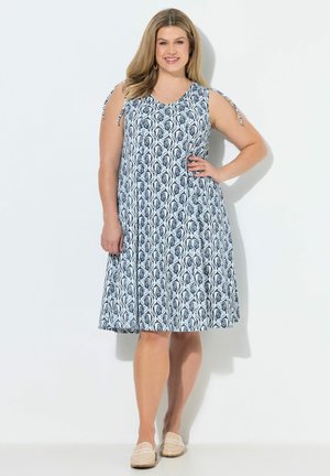 Kobieta w niebiesko-białej, wzorzystej sukience bez rękawów do kolan, z wiązaniami na ramionach, stojąca z jedną ręką na biodrze i uśmiechająca się.