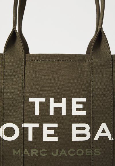 Torebka tote w kolorze oliwkowym z płótna, z białymi literami blokowymi częściowo ukazującymi słowa "THE TOTE BA" oraz "MARC JACOBS" nadrukowanymi poniżej.