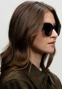 Černé obdélníkové sluneční brýle mají silný rám. Nositelka má dlouhé hnědé vlasy a je oblečená v tmavozelené semišové bundě.